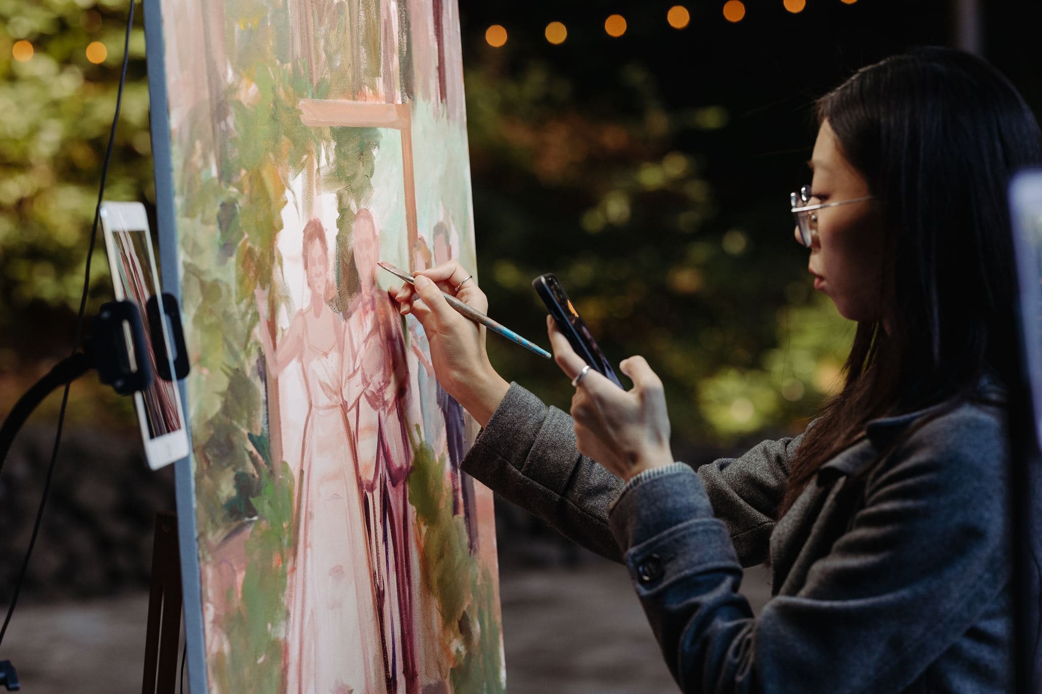 A live wedding painter working on a canvas portrait of the couple in a garden lit with string lights, using an iPad and phone as reference